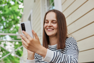 Fototapeta na wymiar Cheerful caucasian attractive adult woman wearing striped shirt using phone sitting on porch of house reading comments in social networks looking at device display.