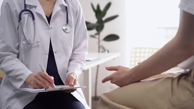 Doctor and patient sitting opposite each other in clinic, closeup. Unknown female physician using clipboard to fill out medical form. Medicine concept