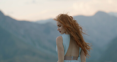 Naklejka premium Carefree caucasian girl with red hair with mysterious glance standing against the wind, it blows her dress and hair. Tranquility, inspiration, freedom concept. Copy space