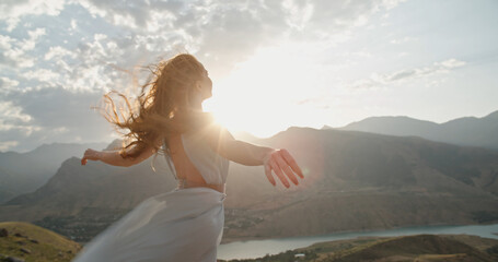 Woman in white dress standing on top of a mountain at sunset with raised hands while wind is...