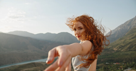 Fototapeta na wymiar Beautiful caucasian girl wearing white dress on top of a mountain. Gorgeous woman looking at camera and reaching her hand out to camera while wind is blowing red hair. Hold my hand and follow me. 