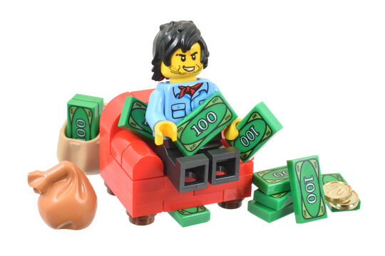 Lego minifigure of happy male is sitting on the chair and counting his money green banknotes in sack. Editorial illustrative image of finance and banking.