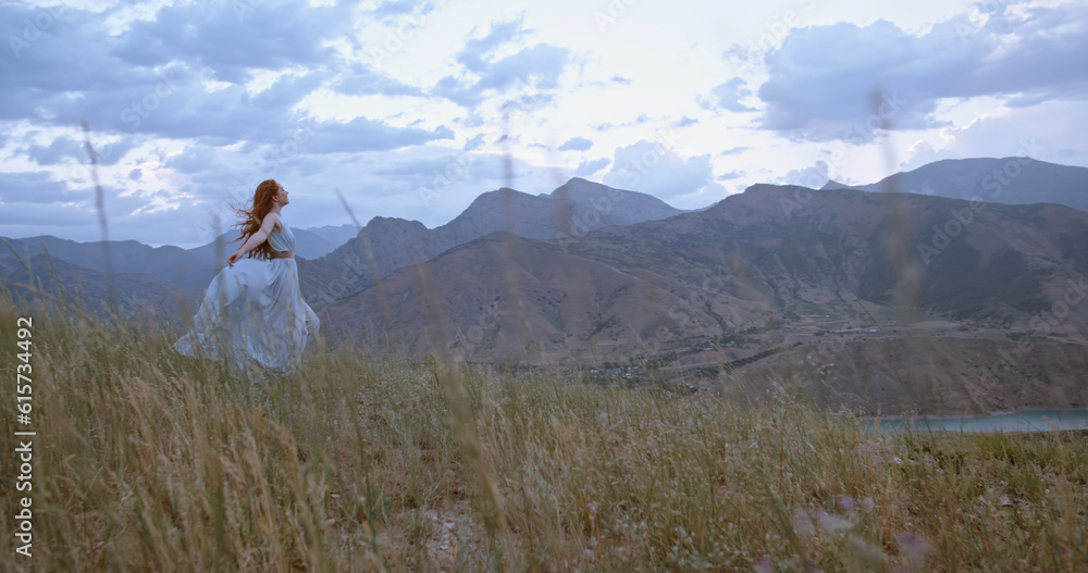 Wall mural young beautiful girl with red hair wearing white dress walking on top of a mountain facing wind blow