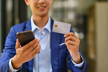 Satisfied asian male worker holding credit card and using smartphone for making orders via iInternet or online payment