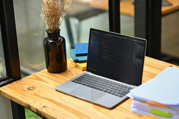 Laptop computer with programming code, document and notepad on wooden table. Web designer...