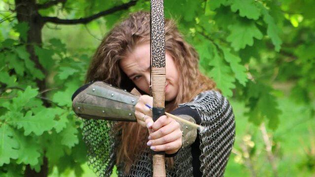 Smooth camera movement, selective focus, portrait of a young woman in chain mail and bracers drawing a bowstring and aiming a bow, against the backdrop of a summer forest.