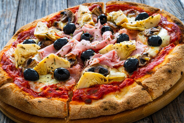 Pizza capricciosa with cooked ham, mozzarella, artichoke and vegetables on wooden table 