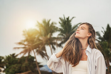 Pretty young woman breathe fresh air on beach at palm trees background. Lovely lady in white shirt relaxing and enjoying vacation in tropical coast. Travel vacation holiday concept. Copy ad text space