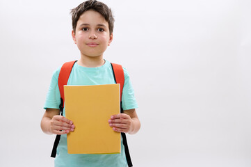 Confident portrait of a Caucasian teenager boy, happy handsome school child 11-13 years old with backpack, holding workbooks and cutely smiling looking at camera, isolated over white studio background