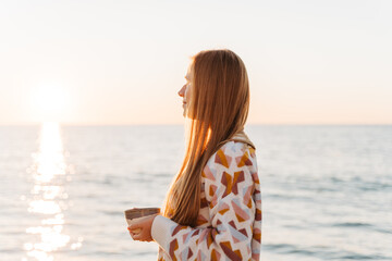 Side portrait of beautiful girl in cozy sweater holding a cup of coffee. Young attractive woman enjoying a view of winter seaside shore with sun trail on the surface of romantic autumn ocean.
