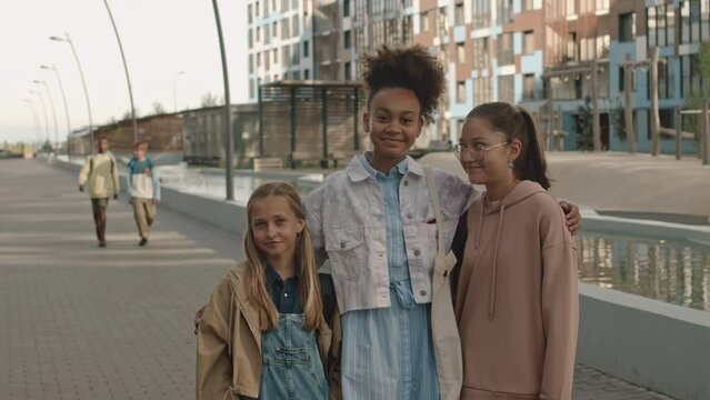 Medium portrait of three multiethnic preteen girls posing for camera standing on pavement on sunny day, walking with classmates after schoolMedium portrait of three multiethnic preteen girls posing fo