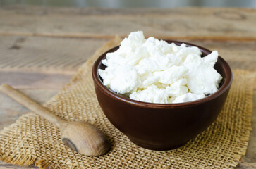 Organic cottage cheese in a brown ceramic bowl with a spoon on burlap on a wooden background. The concept of homemade food. Horizontal orientation.