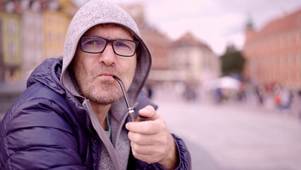 Portrait of adult man with glasses sitting in the hood on square and smoking a tobacco pipe in the...