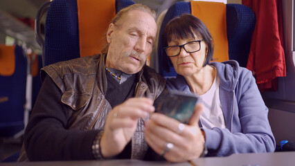 Elderly couple travel by train, the senior is holding a mobile phone in her hand, both are looking at the smartphone and talking with each other