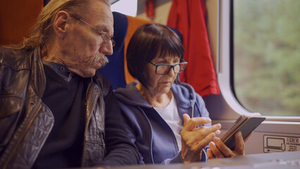 Elderly couple travel by train, the lady is holding a mobile phone in her hand, both are looking at the smartphone and talking with each other