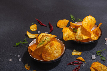 Bowls of homemade potato chips served with chili, parsley, seal salt on black stone background