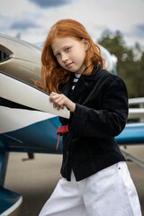 a child girl in white trousers and a black jacket stands near the wing of a small plane