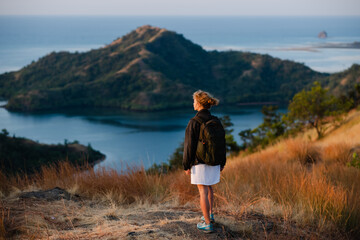 Obraz premium A young girl, blonde, with a backpack, stands on top of a mountain, rear view, overlooking a beautiful panorama of the islands and bays in Indonesia.