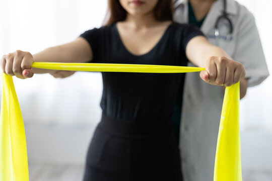 Doctor Practicing Arm And Shoulder Physiotherapy For Female Patient Using Latex Therapy Resistance Band In Exam Room At Hospital. Physical Therapy And Rehabilitation.