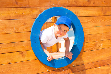 Handsome cheerful happy child boy in a white t-shirt on an eco-friendly wooden playground looks through a round porthole
