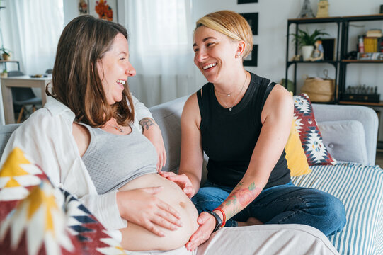 Young woman tender touching partner's female pregnant belly. Same-sex marriage couple on home living room sofa.  Woman's health, happy pregnancy doula supporting and calm mental mood concept image