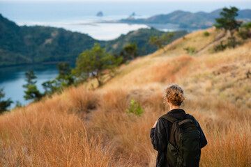 A young girl, blonde, with a backpack, stands on top of a mountain, rear view, overlooking a beautiful panorama of the islands and bays in Indonesia.