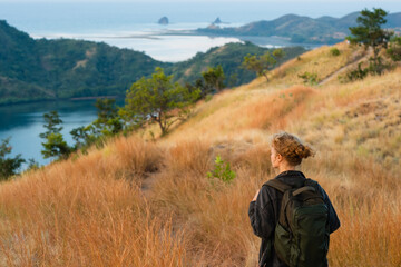 A young girl, blonde, with a backpack, stands on top of a mountain, rear view, overlooking a beautiful panorama of the islands and bays in Indonesia.