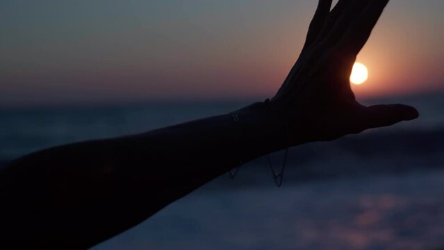 Silhouette of slim female hand holding red sunset sun in circle opening palm closing star. Close-up unrecognizable woman on Mediterranean sea beach at night in darkness