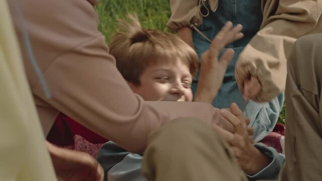 Cropped slowmo of group of schoolkids having fun together while tickling laughing Caucasian boy lying on green grass outdoors in summerCropped slowmo of group of schoolkids having fun together while t