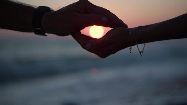 Silhouette of male hand covering female palm holding red sunset sun over Mediterranean sea. Close-up side view unrecognizable loving happy couple enjoying twilight sundown on Cyprus