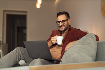 Smiling man laying on the sofa, holding a cup of coffee, using a laptop.