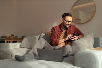 Smiling man checking his social medias, wearing glasses, dressed casually, sitting on the sofa.