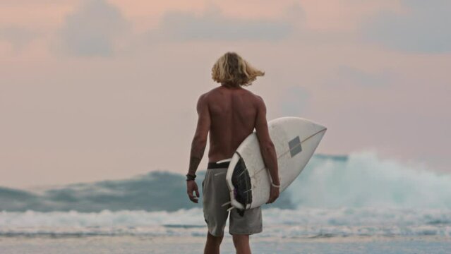Rear view of broad muscular tanned back of male surfer going to sea with raging waves to surf. Concept of weekend evening spent for active recreation, for water sports surfing. Active rest on weekends
