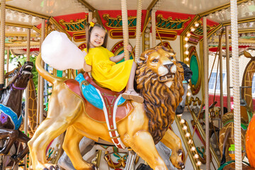 Fototapeta na wymiar Adorable little girl in summer yellow dress at amusement park having a ride on the merry-go-round and eating cotton candy