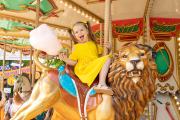 Adorable little girl in summer yellow dress at amusement park having a ride on the merry-go-round and eating cotton candy