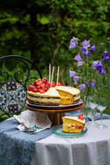 Homemade strawberry Birthday cake with three candles and  slice of cake on glass plate on round table in blooming garden.