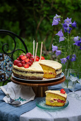 Homemade strawberry Birthday cake with three candles and  slice of cake on glass plate on round table in blooming garden.