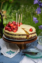 Homemade strawberry Birthday cake with three candles and cut out slice on wooden cake stand on round table in blooming garden.