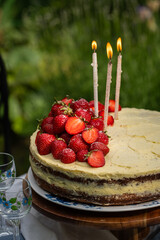 Homemade strawberry Birthday cake  with three lit candles on wooden cake stand on round table in blooming garden.