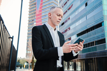 Middle aged businessman using a smartphone to check his stock shares. Well dressed successful business person typing with a cellphone messaging app standing at workplace. Mature executive with phone