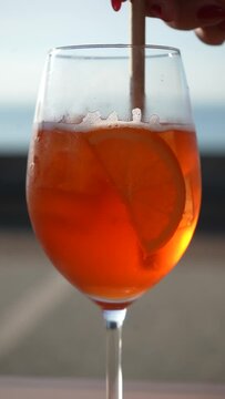 Close-up vertical shot of unrecognizable woman stirring aperol spritz cocktail with straw on background of sunset sky outdoors. Delicious alcoholic drink with citrus fruits in outside summer bar.