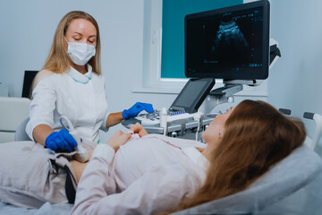 The doctor in a white coat and protective mask examines the patient using an ultrasound scanner....