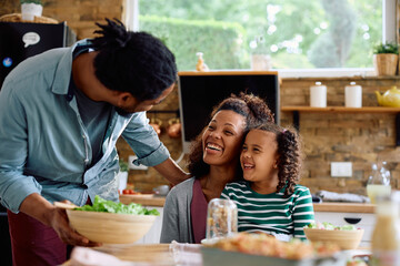 Happy black mother and daughter talking to father who is serving them food at dining table.