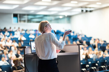 Female speaker giving a talk on corporate business conference. Unrecognizable people in audience at...