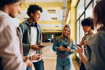Happy teenage girl and her classmates using cell phones in hallway at high school.