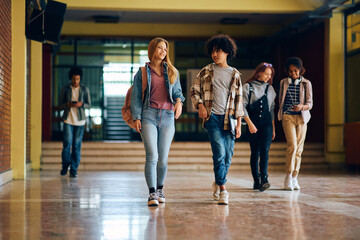 Happy high school friends talk while walking through hallway at school.