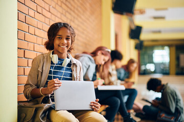 Happy black teenage girl using laptop in hallway at high school and looking at camera.