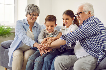 Happy grandparents and little children having fun while spending time together. Cheerful, joyful brother and sister together with grandmother and grandfather sitting on sofa at home and stacking hands