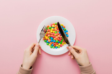 the girl holds cutlery in her hands and eats sweets in a plate. Health and obesity concept, top view on colored background