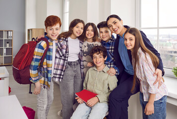 Portrait of cheerful school children and female teacher standing together. Smiling schoolchildren in casual clothes posing with positive teacher in classroom at school